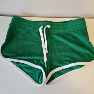 Gröna shorts från H&M - Snygga gröna shorts från H&M med vit snörning och kant. Tillverkade i en mjuk blandning av polyester och bomull, perfekta för en avslappnad stil. De har en sportig look med elastisk midja och kort passform.