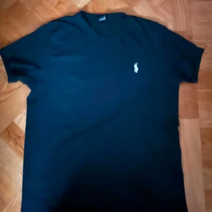 Svart t-shirt från Ralph Lauren - Snygg svart t-shirt från Ralph Lauren med klassisk logga på bröstet. Perfekt för en stilren look. T-shirten har en normal passform och är kortärmad.