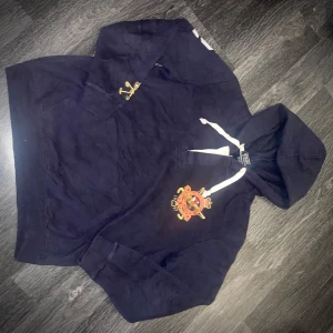 Mörkblå hoodie från Ralph Lauren - Säljer en snygg mörkblå hoodie från Ralph Lauren med broderad logga på bröstet och stort nummer 2 på ryggen, samt har den en logga och nummer 2 på höger ärm. Tröjan har en justerbar huva med vita snören och en praktisk magficka. Perfekt för en avslappnad stil!