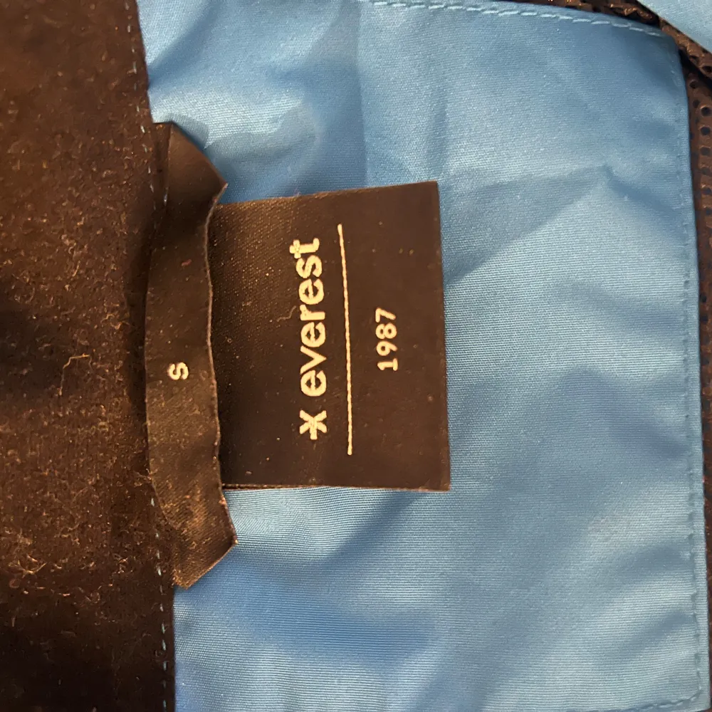 Säljer en blå vindjacka från Everest med huva och dragkedja framtill. Jackan har en stilren design och är perfekt för blåsiga dagar. Den är lätt och smidig att bära med sig.. Takit.