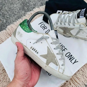 GGDB sneakers med gröna detaljer - Snygga GGDB sneakers i vitt med gröna glittriga detaljer på hälen och en stjärna på sidan. Skorna har grå snörning och en klassisk design med svart sula. Perfekta för en trendig look.