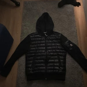 Moncler cardigan - Säljer en snygg cardigan från Moncler med huva och dragkedja. Jackan har långa ärmar och en broderad Moncler-logga på ärmen. Perfekt för kyligare dagar. Storlek L men sitter som M. 