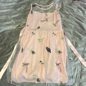 Rosa klänning med broderade blommor - Charmig rosa klänning med tunna axelband och ett lager av transparent tyg med broderade blommor i olika färger. Klänningen har en söt rosett i midjan och ger en romantisk känsla. Perfekt för speciella tillfällen. Perfekt för små flickor 🩷