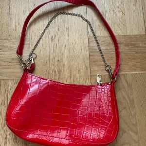 Röd axelväska med krokodilmönster - Säljer en supercool röd axelväska med krokodilmönster. Väskan har en glänsande finish och en kedja som detalj på axelremmen. Den stängs med en dragkedja och är perfekt för att ge din outfit en färgklick. Passar både till vardags och fest!