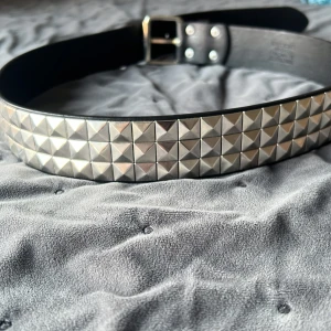 Svart nitat bälte - Säljer ett coolt svart bälte med silverfärgade nitar. Bältet har en klassisk design med en rad av pyramidformade nitar som ger en edgy look. Det är gjort av skinn och har ett robust spänne i silver. Perfekt för att ge lite attityd till din outfit! Aldrig använt 