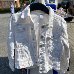 Vit jeansjacka från Lindex - Säljer en snygg vit jeansjacka från Lindex i storlek 140, perfekt för barn mellan 9-10 år. Jackan har en klassisk design med knappar framtill och två bröstfickor. Den är långärmad och gjord i ett slitstarkt material som passar perfekt för vår och sommar.