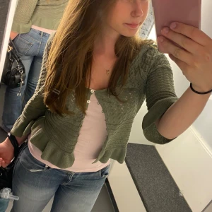 Grön stickad kofta med knappar - Säljer en superfin grön stickad kofta i storlek xs. Den har ett snyggt mönster och knappar framtill. Koftan är croppad och har trekvartsärmar, vilket gör den perfekt för både vår och höst. Den är i nyskick och väldigt bekväm att ha på sig. Perfekt för en casual look eller att klä upp med några accessoarer!