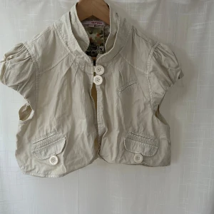 Beige kortärmad väst med knappar - Säljer en superfin beige bolero med korta ärmar och stora knappar framtill. Perfekt för att lägga till en extra touch till din outfit!