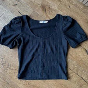 Svart ribbad topp från NA-KD - Säljer en svart ribbad topp från NA-KD. Den har korta puffärmar och knappar framtill, går ej att knäppa upp. Perfekt för hösten!