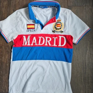 Vit Madrid t-shirt med krage - Säljer en vit t-shirt med krage och tryck från Madrid. Den har en röd och blå rand över bröstet och texten 'MADRID' i vitt. Det finns också en spansk flagga och texten 'SPAIN' på ena sidan samt ett emblem på andra sidan. Perfekt för dig som älskar Spanien!