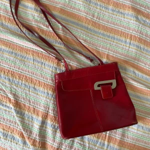 Röd axelväska i skinn - Säljer en snygg röd axelväska i skinn. Väskan har en stilren design med en unik metallspänne på framsidan. Den har flera fack, inklusive ett med dragkedja på baksidan och ett stort huvudfack med dragkedja inuti. Perfekt för både vardag och fest!