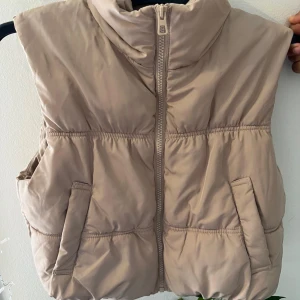 Beige dunväst med dragkedja - Säljer en snygg beige dunväst som är perfekt för höst och vinter. Västen har en dragkedja framtill och två praktiska fickor. Den är i nyskick och passar perfekt för en normal passform. Supermysig och stilren!