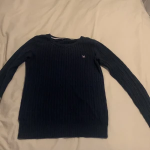 Mörkblå stickad tröja från Gina Tricot - Säljer en snygg mörkblå stickad tröja från Gina Tricot i storlek S. Tröjan har ett diskret kabelstickat mönster och en liten broderad logga på bröstet. Perfekt för höst och vinter, med långa ärmar och ribbade kanter för extra komfort. Passar både till vardags och lite finare tillfällen!