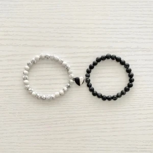 Svart och vitt pärlarmband med hjärtdetalj - Två stilrena armband, ett i svart och ett i vitt, båda med runda pärlor. Det vita armbandet har en svart hjärtdetalj, medan det svarta armbandet har en vit hjärtdetalj. Perfekta för att matcha med olika outfits eller bära tillsammans för en kontrasterande look.