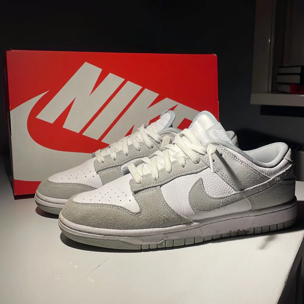 Säljer ett par fräscha Nike Dunk sneakers i vitt och grått. De är i nytt skick och kommer med originalkartong. Perfekta för både vardag och träning. Skorna har en snygg kombination av skinn och mocka med klassisk snörning.. Skor.