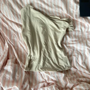 Beige oneshoulder topp - Säljer en stilren beige oneshoulder topp i mjukt material. Lätt att matcha med olika outfits. 