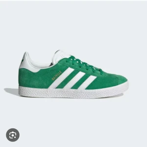 Gröna Adidas Gazelle sneakers - Säljer ett par snygga gröna Adidas Gazelle sneakers i mocka. De har de klassiska vita ränderna och en vit sula. Perfekta för en casual look och passar till nästan allt. Skorna är i nytt skick och kommer med originalkartong. Perfekt för dig som vill ha en stilren och tidlös sneaker! 💚