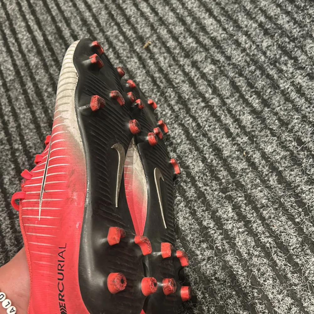 Säljer ett par Nike Mercurial fotbollsskor i rött och vitt med svarta detaljer. Skorna har snörning och är i bra skick, perfekta för fotbollsträning eller match. De har en snygg design med Nike-loggan på sidan och dobbar för bra grepp på planen.. Skor.