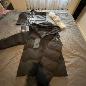 Canada Goose Wyndham Parka - Säljer en svart dunjacka från Canada Goose i storlek L med black label. Jackan är perfekt för kalla vinterdagar med sin tjocka vaddering och avtagbar pälskrage. Den har en dragkedja och knappar framtill samt en broderad logga på ärmen. Jackan är i nyskick och kommer med originalpåse.
