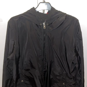 Svart vindjacka från Moncler - Säljer en svart vindjacka från Moncler i mycket bra skick. Jackan har en dragkedja framtill och en huva med ränder i rött, vitt och blått. Perfekt för höst och vår, och den är långärmad. Moncler-loggan finns på ärmen. NFT funkar även på denna modellen!