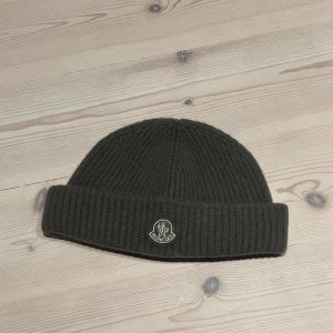 Olivgrön  mössa  Moncler - Säljer en snygg olivgrön mössa från Moncler i ribbat material. Perfekt för kyliga dagar med sin stilrena design och varma känsla. Den har en uppvikt kant och Moncler-loggan framtill. Passar både till vardags och mer uppklädda tillfällen.
