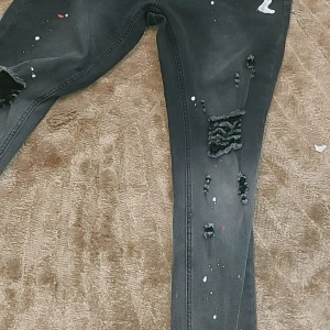 Svarta jeans med slitningar och färgstänk - Säljer ett par svarta jeans med coola slitningar och färgstänk i vitt och rött. De har en tight passform och är perfekta för en avslappnad och trendig look. Jeansen är i välanvänt skick men har fortfarande mycket att ge. Perfekta för höst och vår! Passar xs och xxs