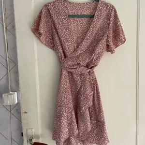 Rosa blommig klänning från Shein - Säljer en superfin rosa blommig klänning från Shein. Den har en omlott-design med ett knytband i midjan och korta ärmar. Perfekt för sommaren eller en fest! Klänningen är i nyskick och väldigt bekväm att ha på sig.