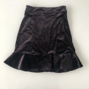 Svart sammetskjol med volang - Säljer en superfin svart sammetskjol med volangdetalj längst ner. Kjolen har en dragkedja bak och är i ett mjukt och glansigt material. Perfekt för fest eller en utekväll! 🖤