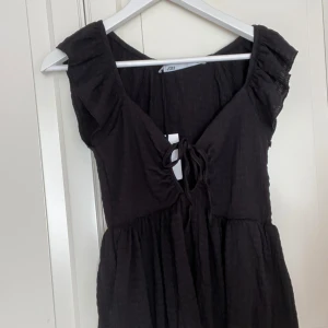 Svart topp med knytning - Säljer en svart topp med knytning framtill. Toppen är helt oanvänd med prislappen kvar, volangdetaljer vid axlarna. Perfekt för både vardag och fest!