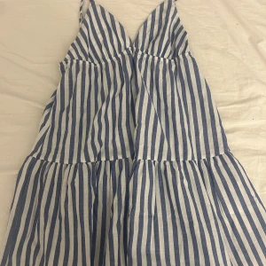 Randig sommarklänning från Shein - Säljer en superfin randig sommarklänning från Shein i storlek M. Klänningen är vit och blå med smala axelband och en v-ringad hals. Den är tillverkad av 65% bomull och 35% polyester, vilket gör den både bekväm och luftig. Säljer då den inte används (prislapp är kvar)