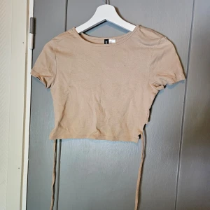 Beige croppad topp från H&M Divided - Säljer en beige croppad topp från H&M Divided. Toppen är kortärmad och har en tight passform. lite kortare i bak för att man ska knyta snörena för en fin detalj. knappt använd