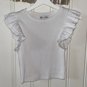 Vit ribbad topp från Zara - Säljer en superfin vit ribbad topp från Zara. Den har volangdetaljer på ärmarna. Toppen är i storlek Xss-Xs och är i nyskick.