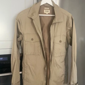 Beige jacka från East West - Säljer en snygg beige jacka från East West. Den har en klassisk design med två bröstfickor med knappar och en dragkedja framtill. Jackan är i storlek herr M och passar perfekt för vår och höst. Den är i bra skick och har en normal passform. Perfekt för en casual look!