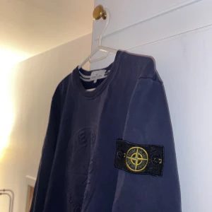 Mörkblå tröja från Stone Island - Säljer en snygg mörkblå tröja från Stone Island i storlek XL. Tröjan har en klassisk rund hals och en cool patch med logga på vänster ärm. Perfekt för höst och vinter, den är både stilren och bekväm. Passar bra till både vardag och fest!