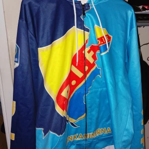 Blå och gul jacka med DIF-logga - Säljer en blå och gul jacka med stor DIF-logga på framsidan. Jackan har dragkedja och huva med vita snören. Perfekt för att visa ditt stöd för laget! Jackan är i bra skick och har texten 'Järnkaminerna' på framsidan.