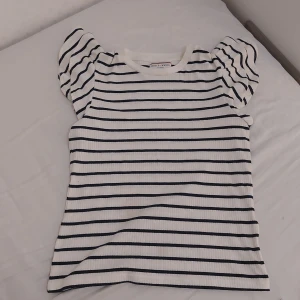 Randig topp från Holly & Whyte - Säljer en superfin randig topp från Holly & Whyte by Lindex. Den är vit med svarta ränder och har korta puffärmar som ger en söt touch. Toppen är ribbad och gjord i bomull, vilket gör den både bekväm och stilren. Perfekt för både vardag och fest!