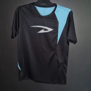 Svart och blå t-shirt - Säljer en snygg svart och blå t-shirt från D-way. Den har en cool design med blåa detaljer på axeln och sidan. Perfekt för träning eller vardagsbruk. T-shirten är gjord av polyester och har en normal passform. Den är i nyskick och redo för en ny ägare!