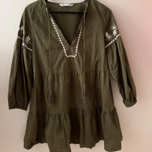 Olivgrön klänning från Zara - Säljer en superfin olivgrön klänning från Zara. Den har en v-ringad hals och trekvartsärmar med vackra broderade blommor i vitt. Klänningen har en lös passform och volanger nedtill. Perfekt för sommaren och hösten! 🌸