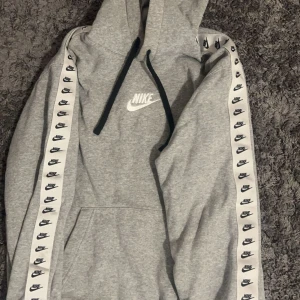 Grå hoodie från Nike - Säljer en grå hoodie från Nike i storlek M. Den är superbekväm och perfekt för höst och vinter. Hoodien har en stor ficka på magen och en justerbar huva med svarta snören. På ärmarna finns det vita band med små Nike-loggor. Pris kan diskuteras  