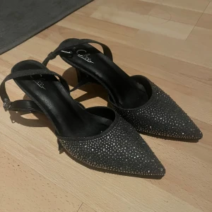 Svarta-grå glittriga pumps med spetsig tå - Snygga svarta pumps med glittrig yta och spetsig tå. Perfekta för en kväll ute eller en speciell tillställning. Skorna har en elegant design med en smal rem runt ankeln.