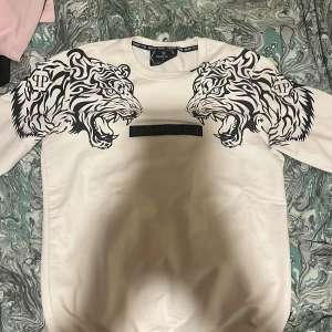 Vit tröja från Philipp Plein med tigertryck - Säljer en vit tröja från Philipp Plein med coolt tigertryck i svart på bröstet. Tröjan har rund hals och är i en stilren design med märkets logga på bröstet och i nacken. Perfekt för dig som gillar att sticka ut med stil!