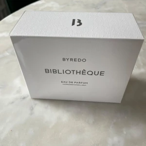 Byredo Bibliothèque Eau de Parfum 100ml - Säljer en elegant Byredo Bibliothèque Eau de Parfum i en stilren vit förpackning. Denna doft fångar essensen av en klassisk biblioteksmiljö med noter av trä och läder. Perfekt för den som älskar sofistikerade och tidlösa dofter.