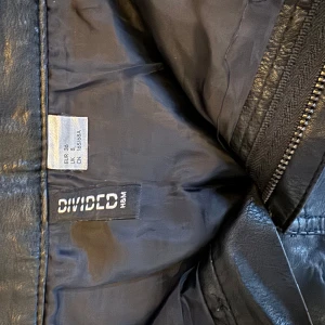 Svart skinnkjol från H&M Divided - Snygg svart skinnkjol från H&M Divided med dragkedja och knapp framtill. Kjolen har praktiska fickor både fram och bak. 