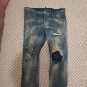 Blå jeans från Dsquared2 - Säljer ett par blå jeans från Dsquared2 med slitna detaljer och färgstänk. Jeansen har en klassisk femficksdesign och knappgylf. Perfekta för en avslappnad och trendig look.