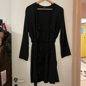 Svart långärmad kappa från Gina Tricot - Säljer en stilren svart kappa från Gina Tricot i storlek 34. Den har långa ärmar och ett knytband i midjan för en snygg passform. Perfekt för att komplettera din outfit med en elegant touch. Lite sönder vid armen 
