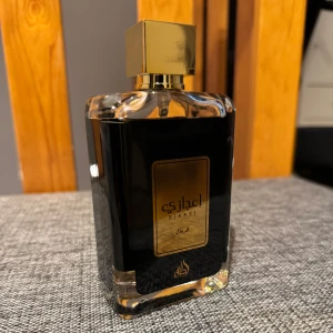 Lattafa Ejaazi Gold EDP - Elegant parfymflaska från Lattafa med en lyxig svart och guld design. Flaskan har en fyrkantig form med en gyllene kork som ger en exklusiv känsla. Perfekt för den som söker en sofistikerad doftupplevelse. Ca 90ml kvar av 100. Lådan medföljer ej.