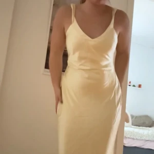 Ljusgul långklänning med öppen rygg - Elegant ljusgul långklänning med smala axelband och en v-ringad framdel. Klänningen har en stilfull öppen rygg med korsade band, vilket ger en extra touch av elegans. Perfekt för sommarens festligheter!