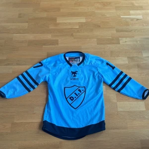 Blå hockeytröja från CCM med DIF-logga - Säljer en blå hockeytröja från CCM med DIF-logga på framsidan. Tröjan har långa ärmar med mörkblå ränder och nummer 17 på både ärmar och rygg. Perfekt för unga fans av Stockholms stolthet! Tröjan är även helt oanvänd