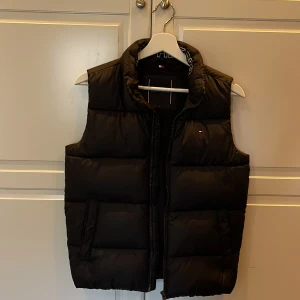 Svart dunväst från Tommy Hilfiger - Snygg svart dunväst från Tommy Hilfiger med dragkedja och hög krage. Diskret logotyp på bröstet och märkesnamn på insidan av kragen.
