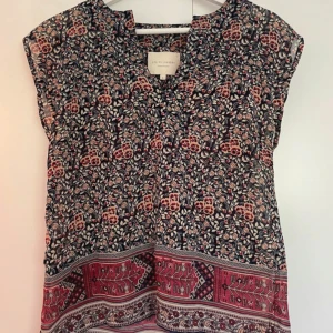 Blommig blus från Lollys Laundry - Säljer en vacker blommig blus från Lollys Laundry i storlek S. Blusen har ett färgglatt mönster med inslag av rött och beige på en mörkblå bas. Den är kortärmad och har en avslappnad passform.